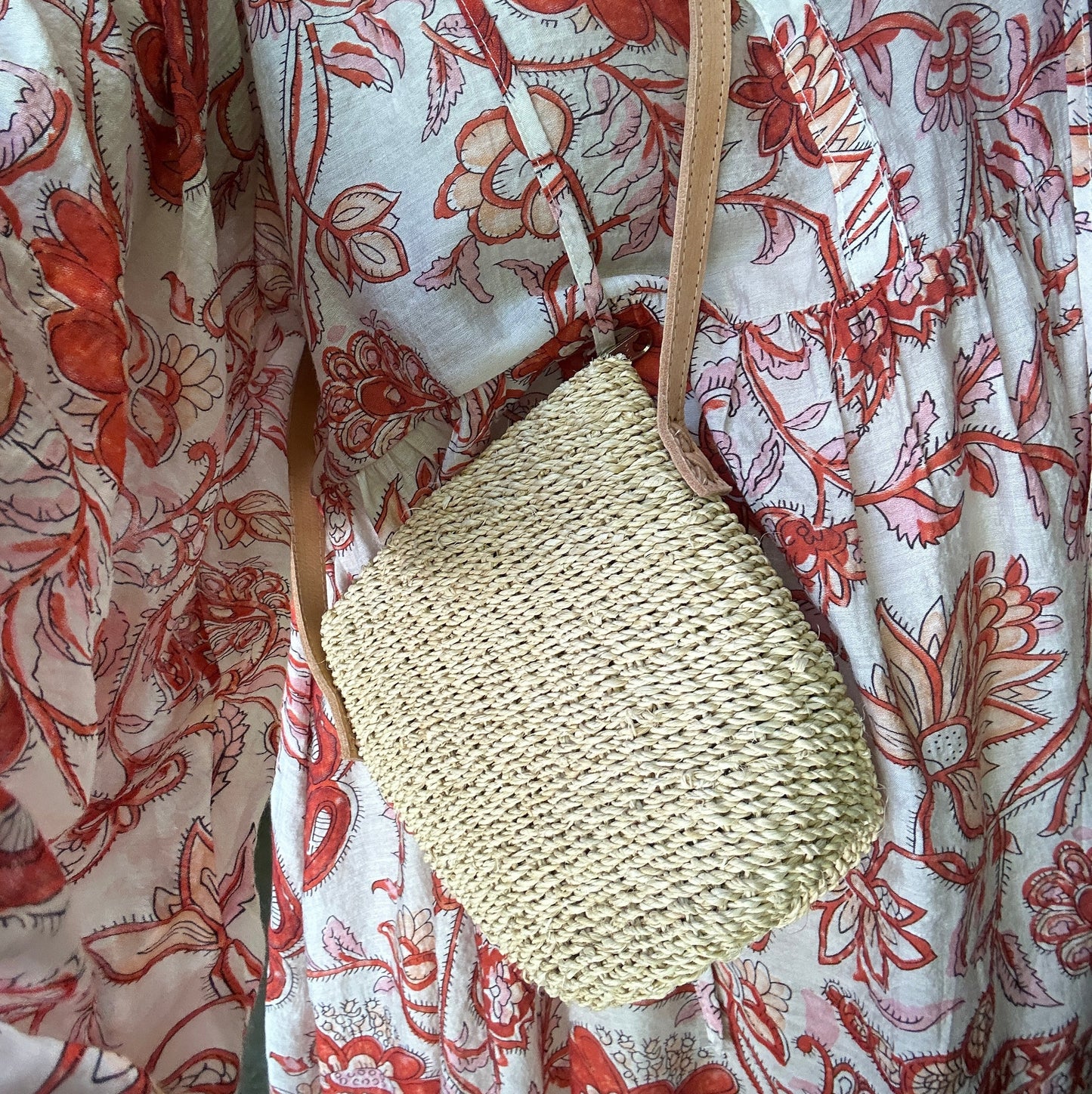 A woven straw shoulder bag with a genuine leather tan strap, shown worn over a white dress with an orange floral print.