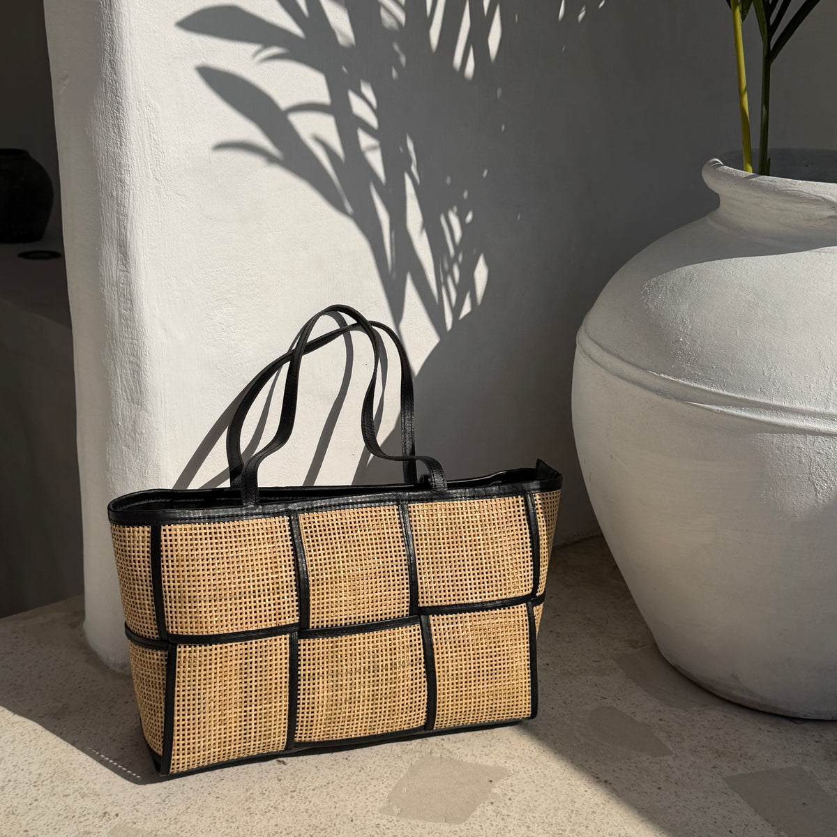A woven rattan tote bag with black trim and handles, styled on a stone floor next to a large white ceramic pot with leafy shadows cast on a white wall.