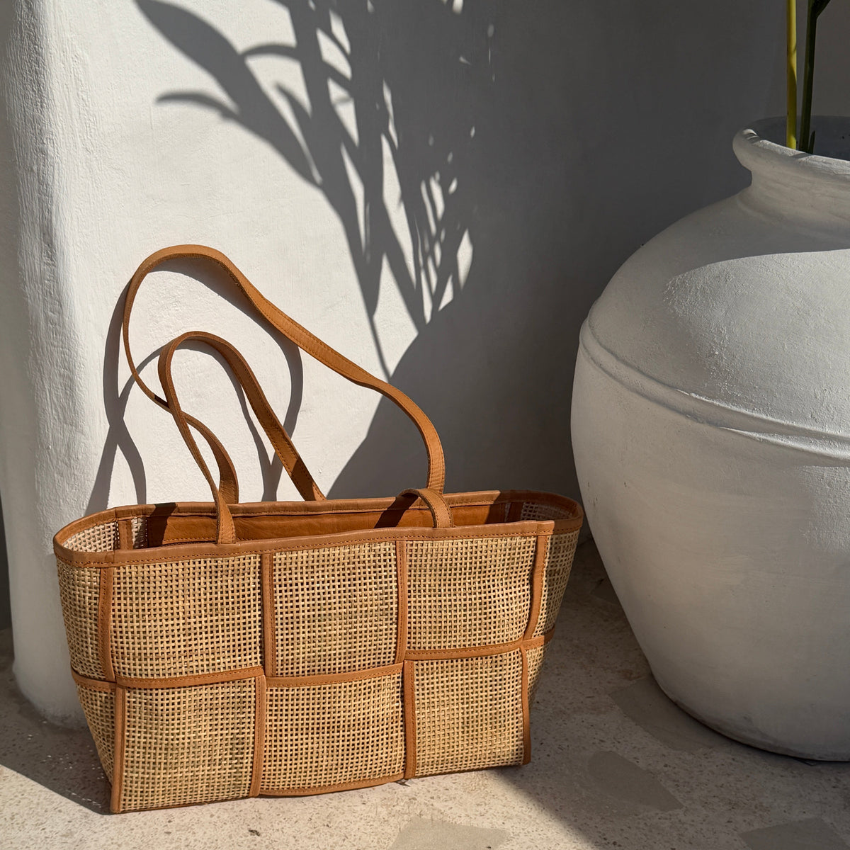 A woven natural fiber tote bag with tan leather handles and trim, styled on a stone floor next to a large white ceramic pot with leaf shadows cast on a white wall.