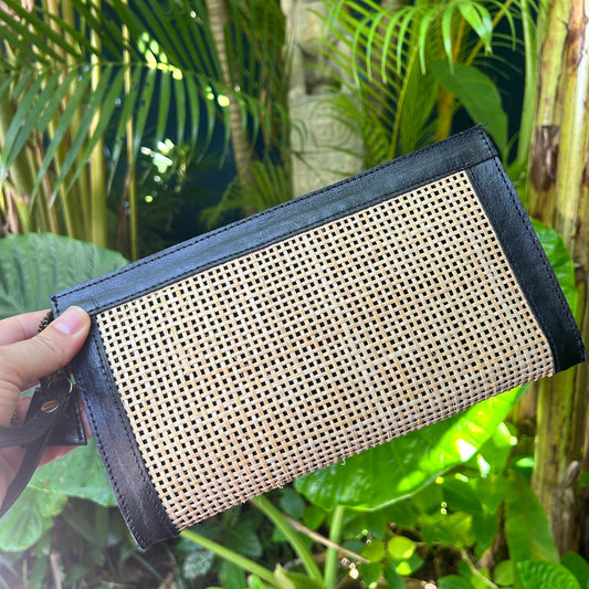 Black genuine leather and rattan clutch bag with wrist strap and zipper - being held in front of tropical greenery