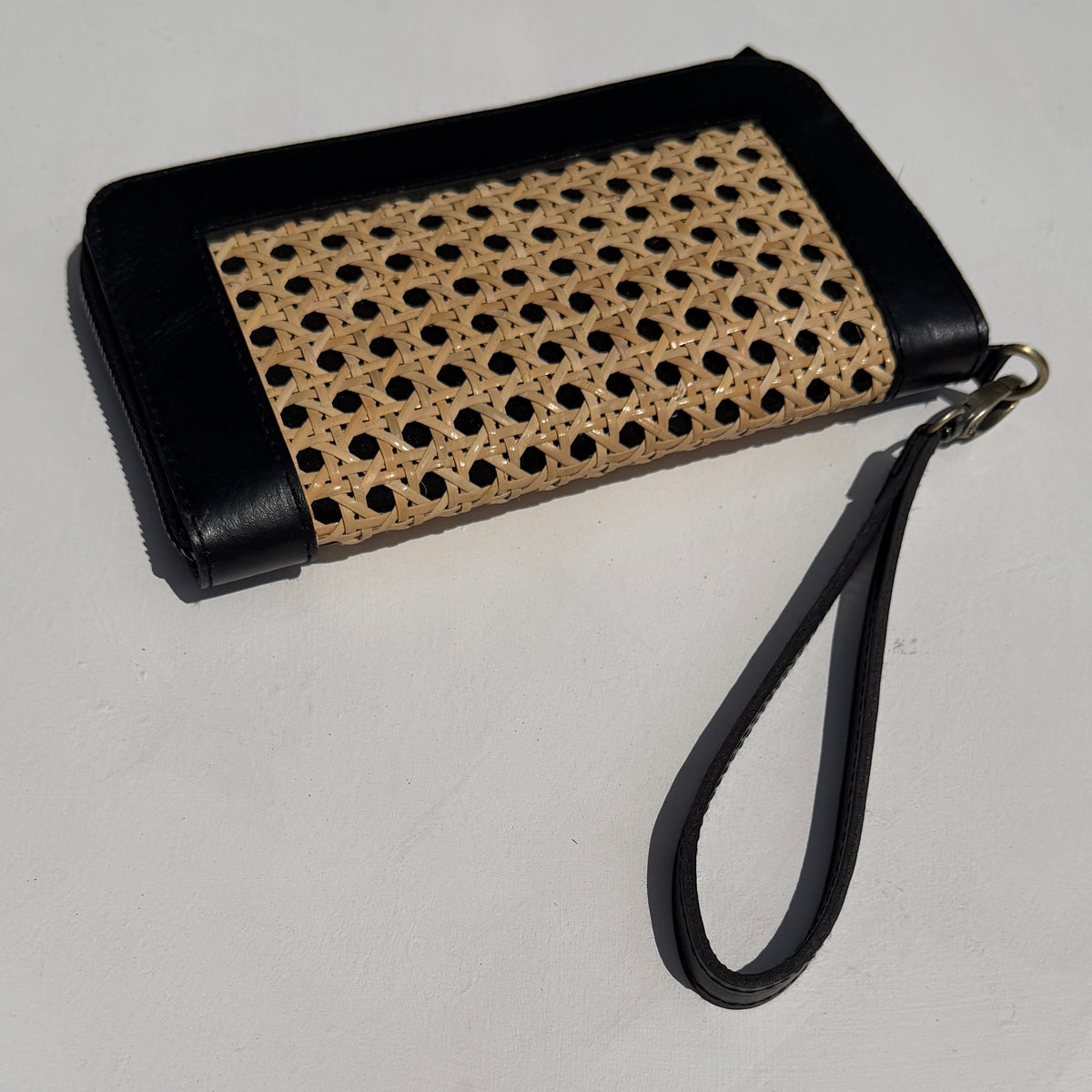 A black leather wristlet clutch with a natural woven cane panel on the front and a matching black wrist strap, shown on a light gray background.