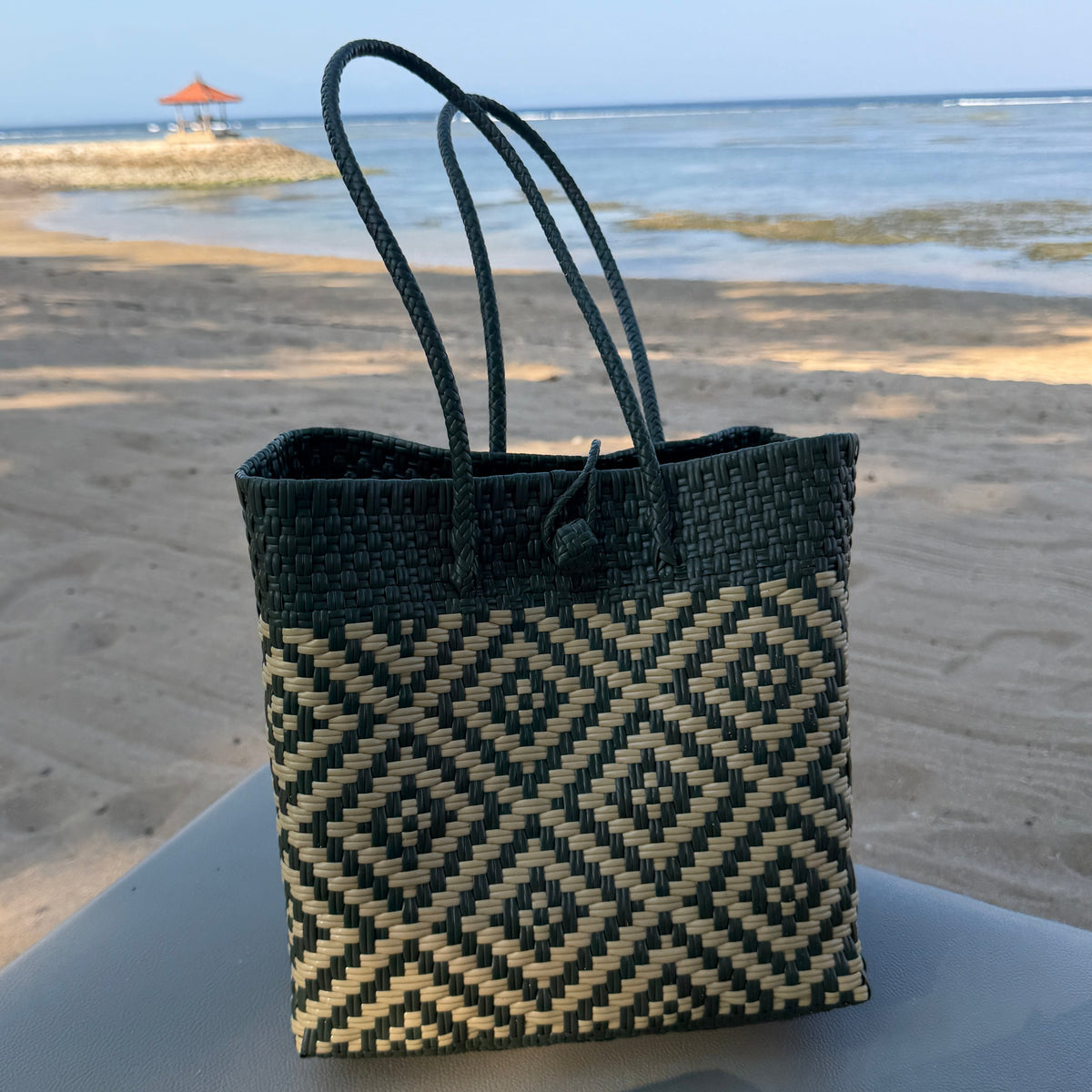 A woven tote bag with a dark green and beige geometric diamond pattern sits on a sunbned on a sandy beach with the ocean in the background.