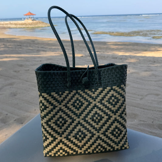 A woven tote bag with a dark green and beige geometric diamond pattern sits on a sunbned on a sandy beach with the ocean in the background.