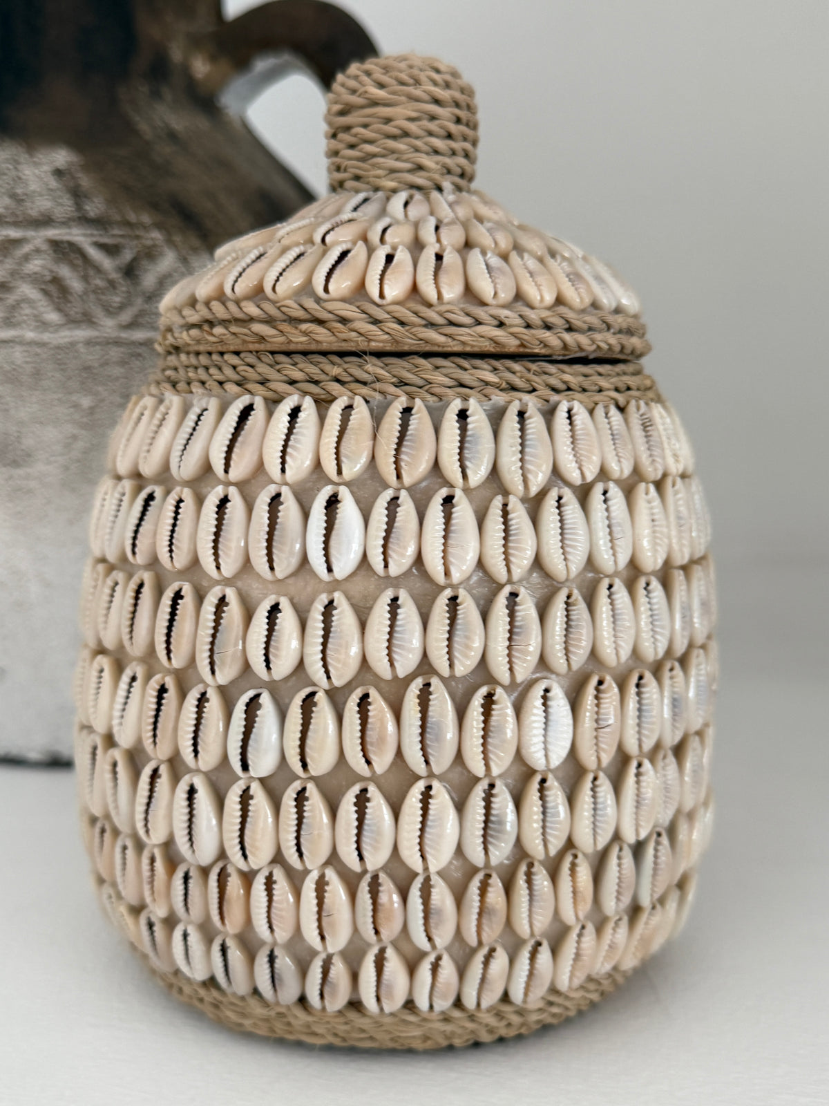 A lidded storage jar decorated with rows of cowrie shells and wrapped in rope, shown against a neutral gray background.