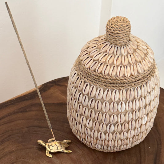 A decorative woven basket with a lid, covered in small cowrie shells and tied with rope, displayed on a wooden surface next to a brass turtle incense holder and incense stick.
