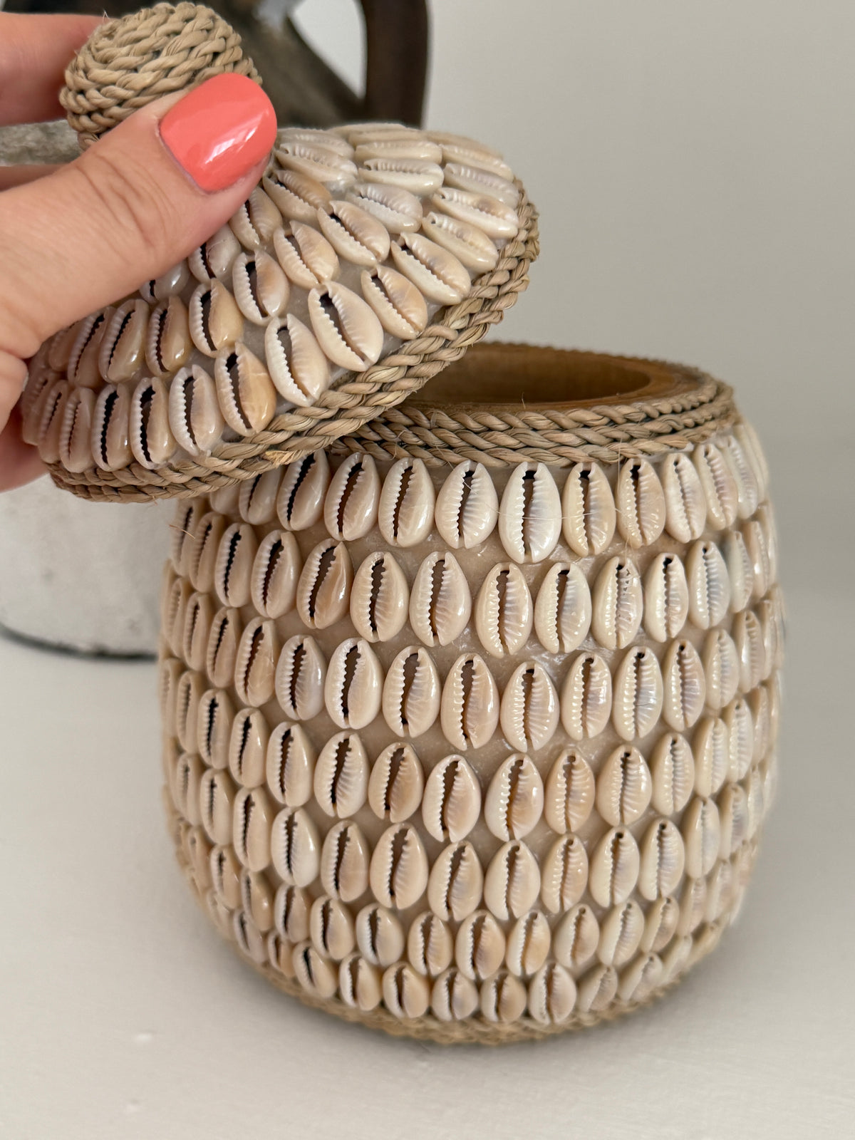 A hand with coral nail polish holds the lid of a woven storage jar covered in beige cowrie shells, set against a neutral light gray background.