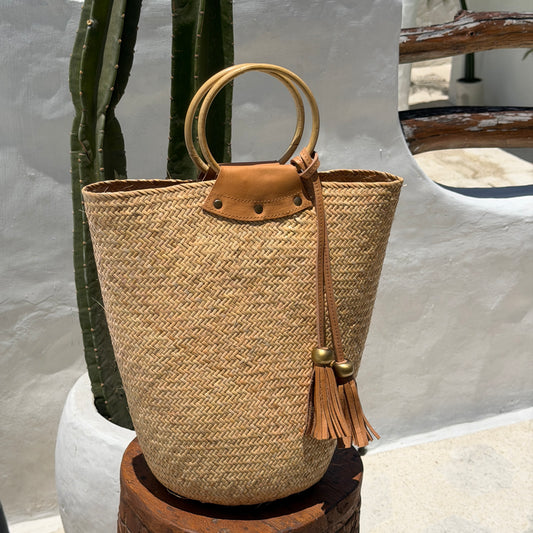 A woven straw bucket bag with circular wooden handles and a tan leather tassel detail, displayed on a wooden stool against a white stucco wall with a cactus in the background.
