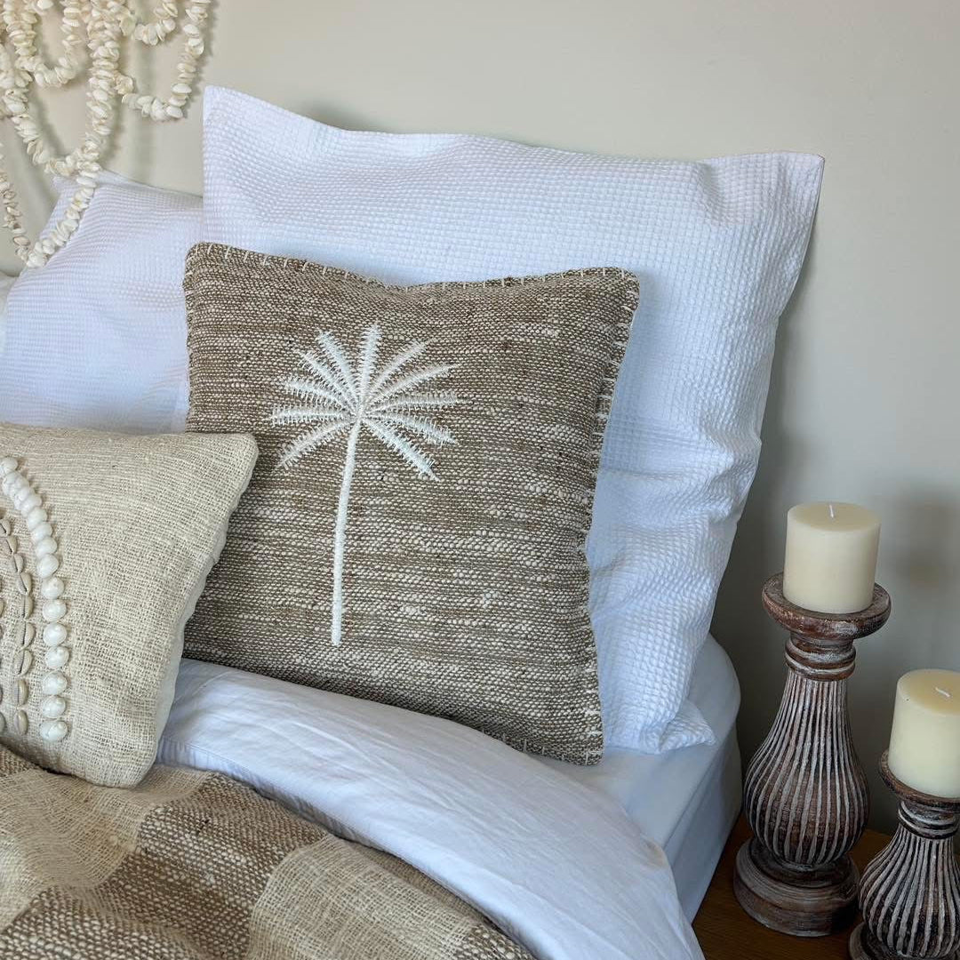Mushroom colour cushion cover with embroidered white palm tree motif 40cm x 40cm.  Displayed on a bed with larger white cushion on background and candle sticks beside.  A shell wall hanger is above the bed.