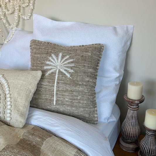 Mushroom colour cushion cover with embroidered white palm tree motif 40cm x 40cm.  Displayed on a bed with larger white cushion on background and candle sticks beside.  A shell wall hanger is above the bed.