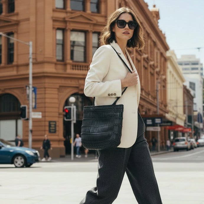 A woman walking on a city street wearing a cream blazer, dark trousers, and sunglasses, carrying a large black leather woven tote bag.