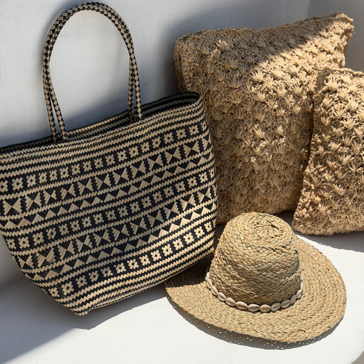 A woven tote bag with a black and beige geometric pattern, styled alongside two textured straw pillows and a matching straw hat on a white surface with natural sunlight.