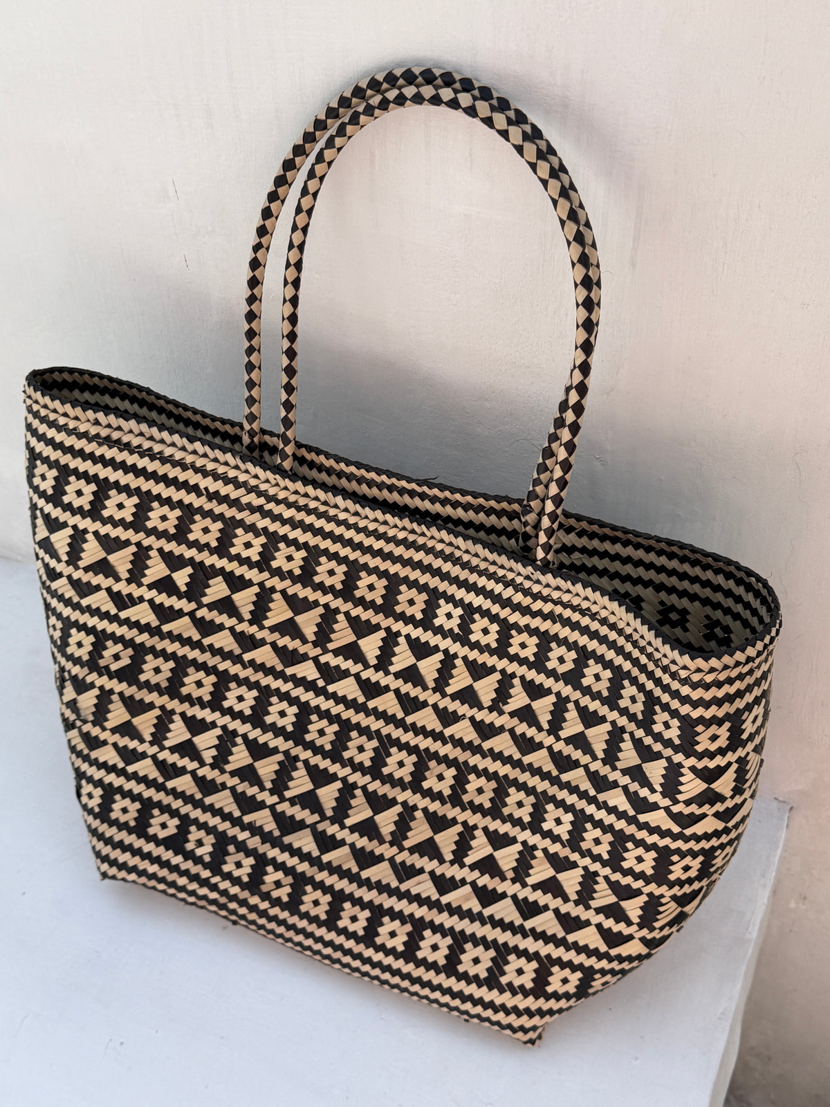 A woven straw tote bag with a black and natural beige geometric pattern, featuring two top handles, set against a plain white wall.