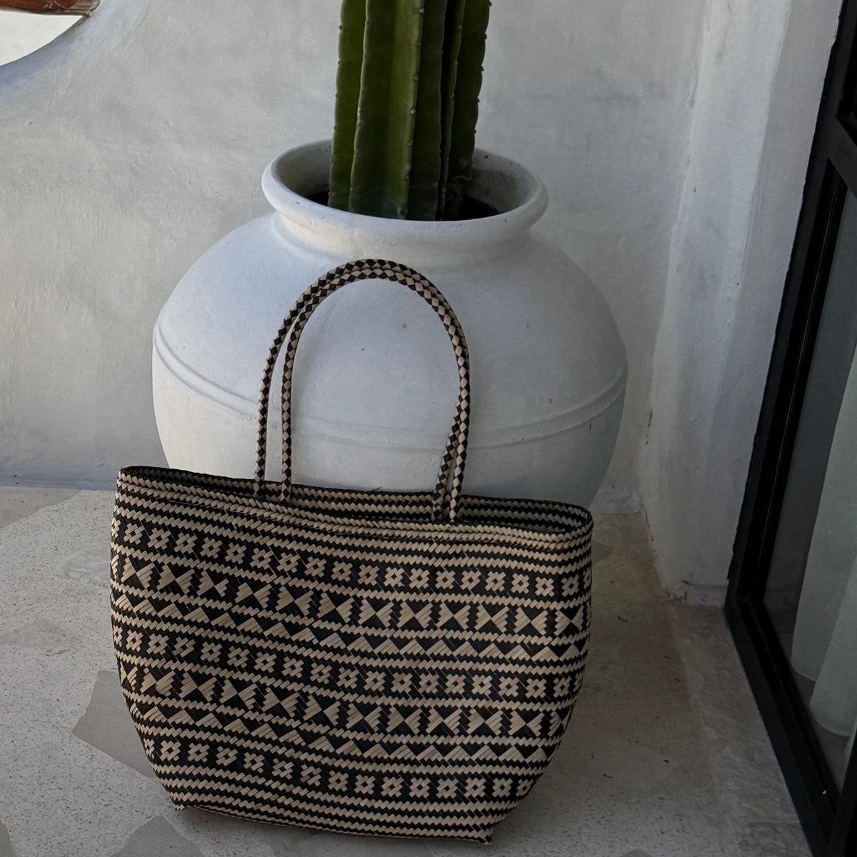 A woven tote bag with a black and beige geometric pattern, featuring two top handles, displayed in front of a large white ceramic planter with a green cactus.