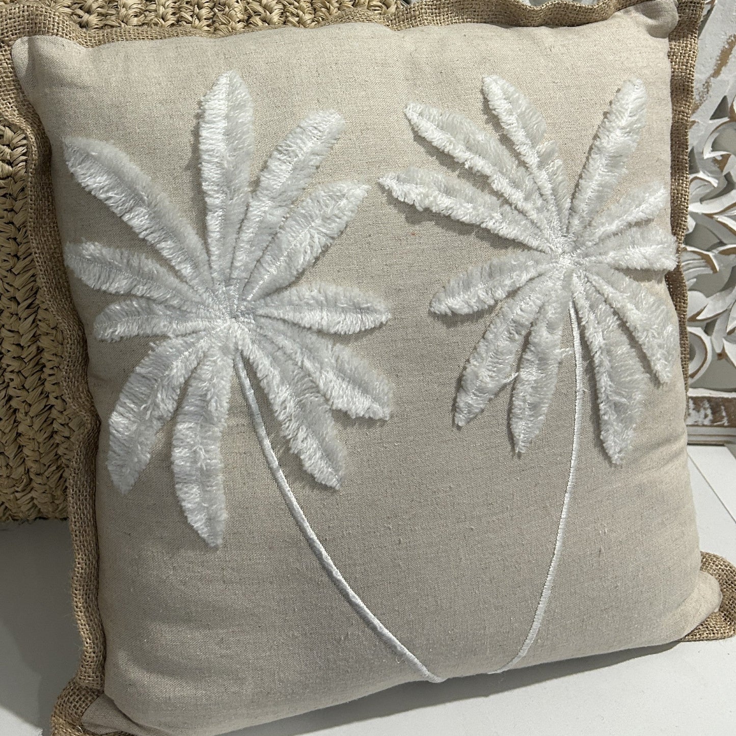 A square beige linen throw pillow with two large white embroidered palm tree designs, styled on a woven straw cushion in front of a white carved wooden screen.