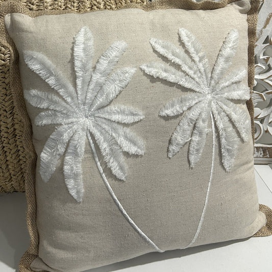 A square beige linen throw pillow with two large white embroidered palm tree designs, styled on a woven straw cushion in front of a white carved wooden screen.