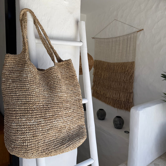 A large woven straw tote bag with two shoulder straps hangs on a white ladder in a bright, modern room with white walls and boho decor.