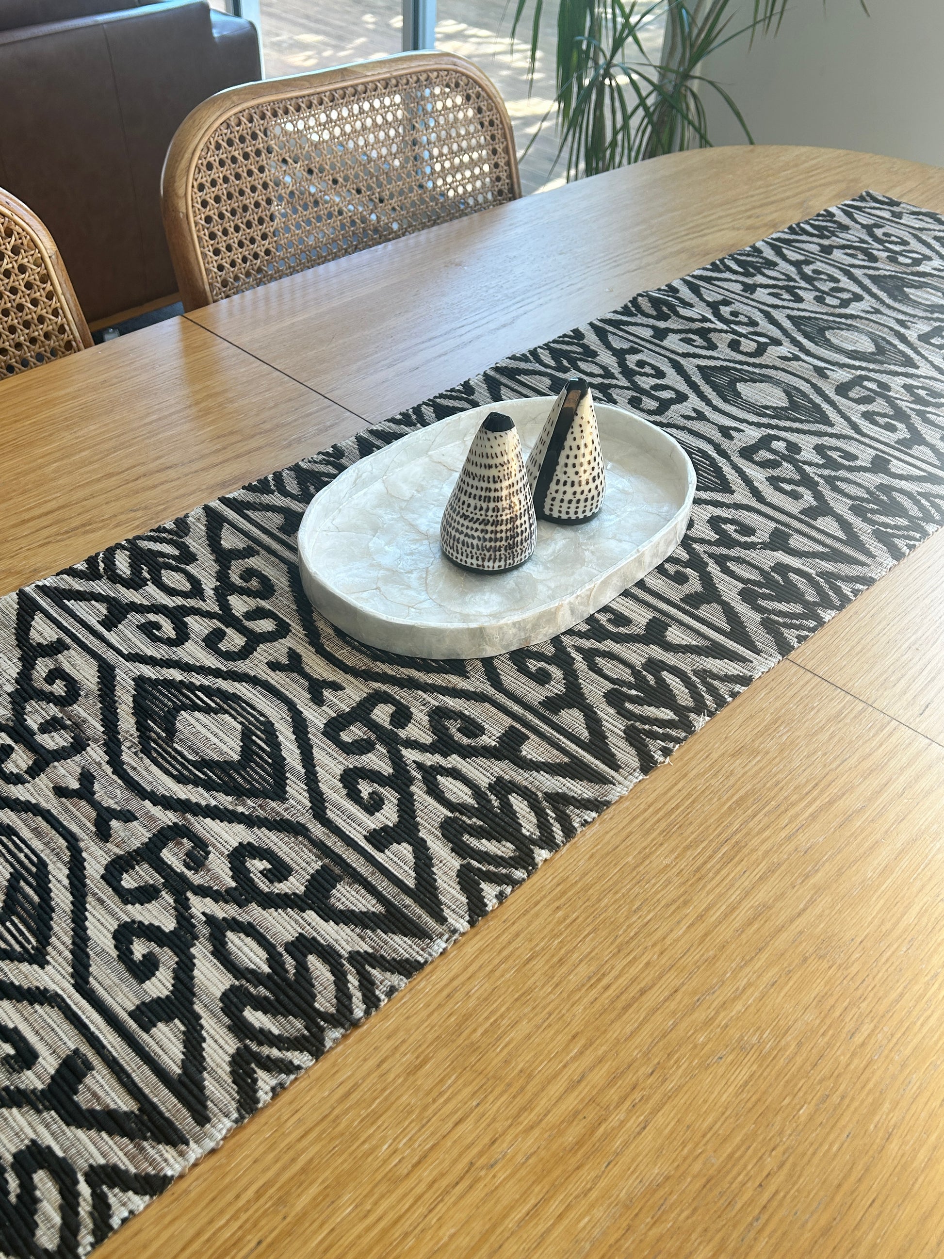 Shell salt and pepper shakers on a capiz shell tray.  Sat on a black and natural table runner atop an oak table with rattan dining chairs. Coastal luxe