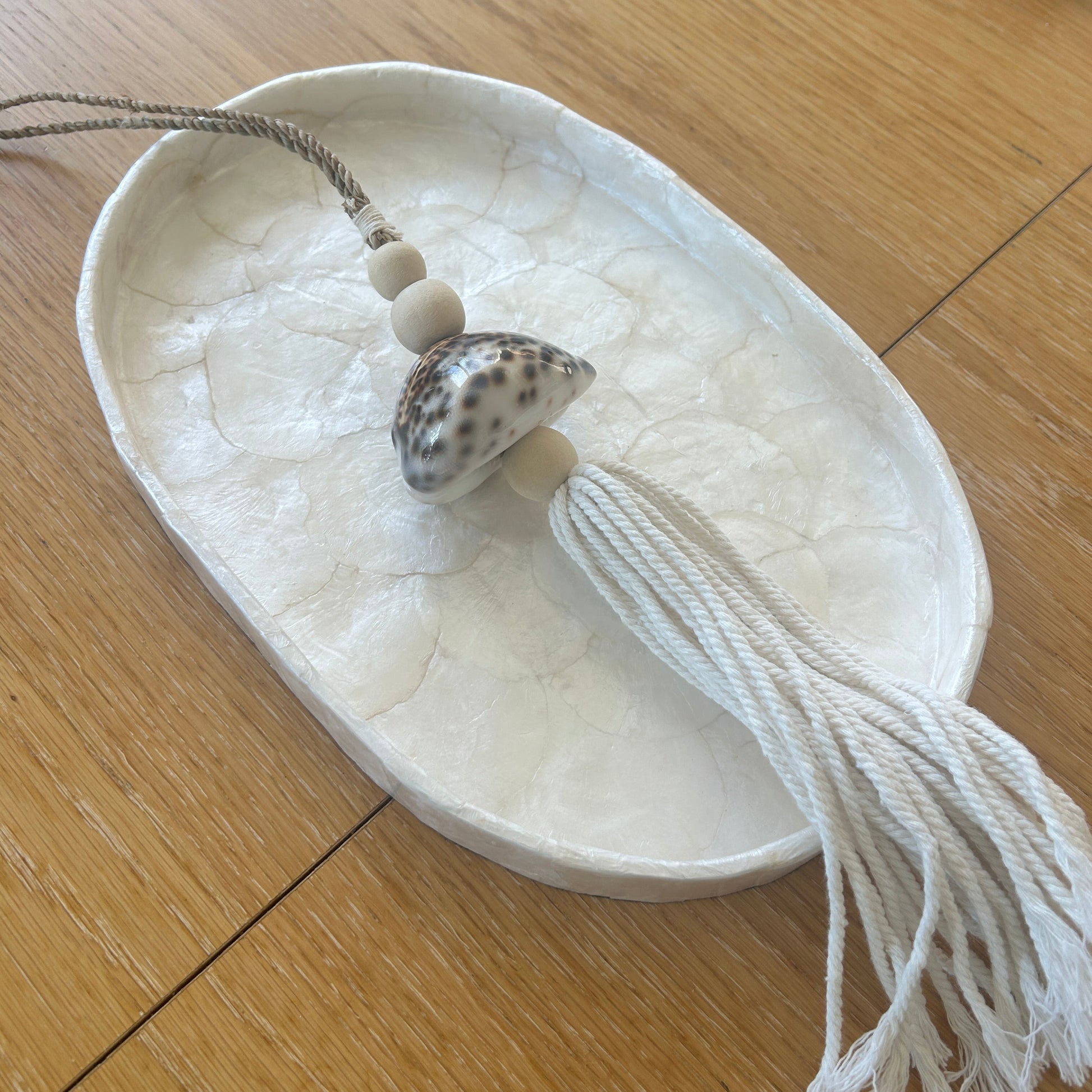 Small shell and decorative item on a white oval dish with tassels on a wooden table.