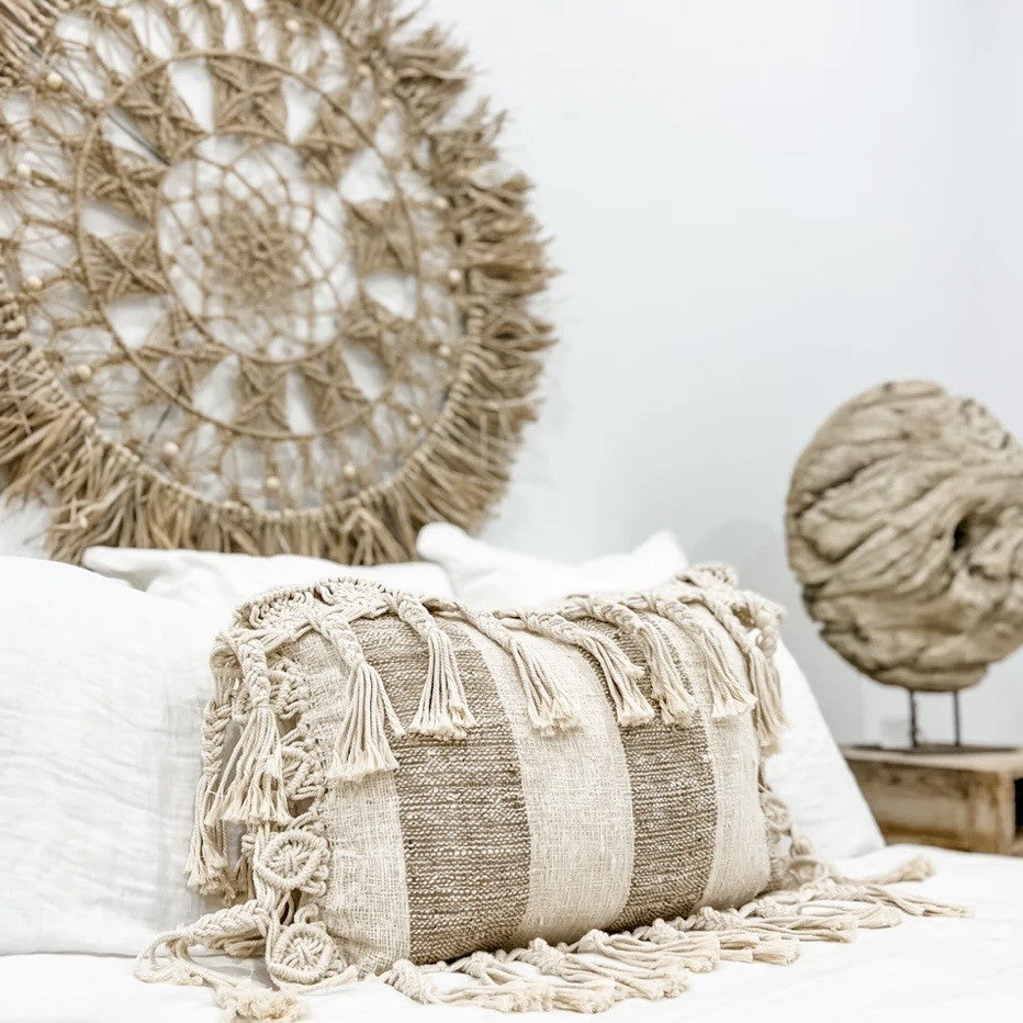Natural and mushroom colour striped decorative pillow with tassels - on a white surface, with a woven wall art piece in the background.