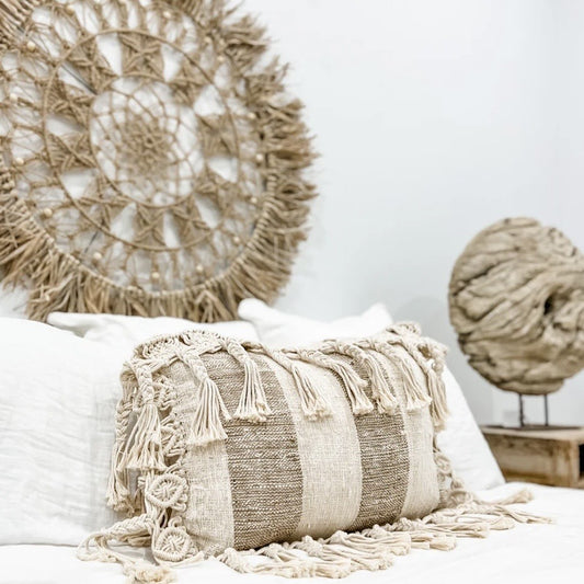 Natural and mushroom colour striped decorative pillow with tassels - on a white surface, with a woven wall art piece in the background.