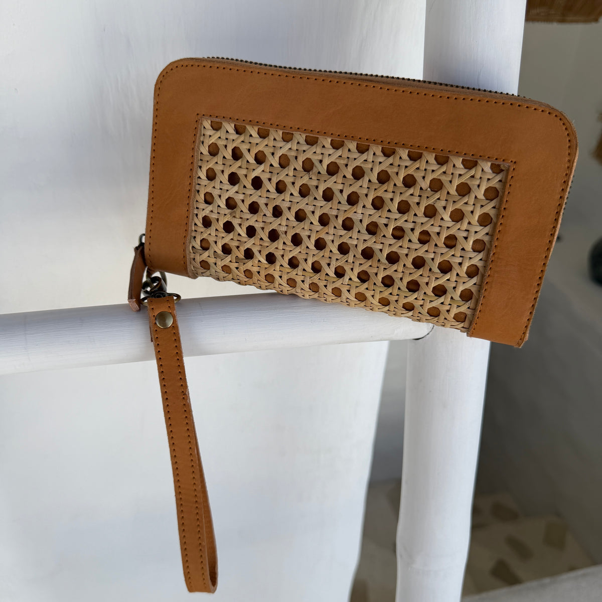A tan leather zip-around wallet with a woven rattan panel on the front and a wrist strap, resting on a white chair.
