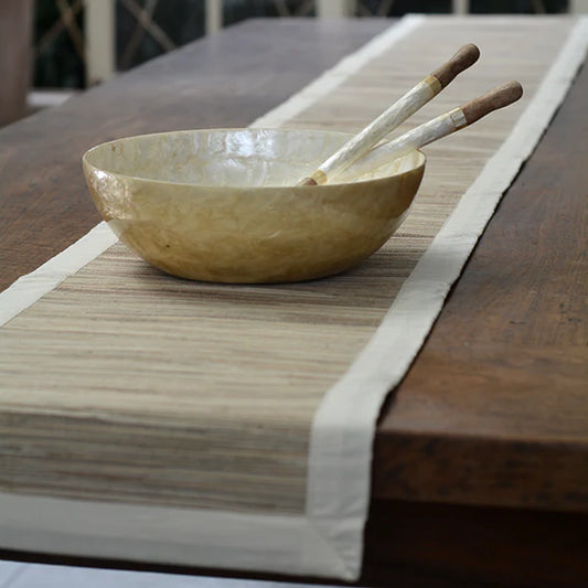 A beige and brown table runner made of water hyacinth with Balinese cotton endek, displayed on a wooden dining table with a bowl and utensils.