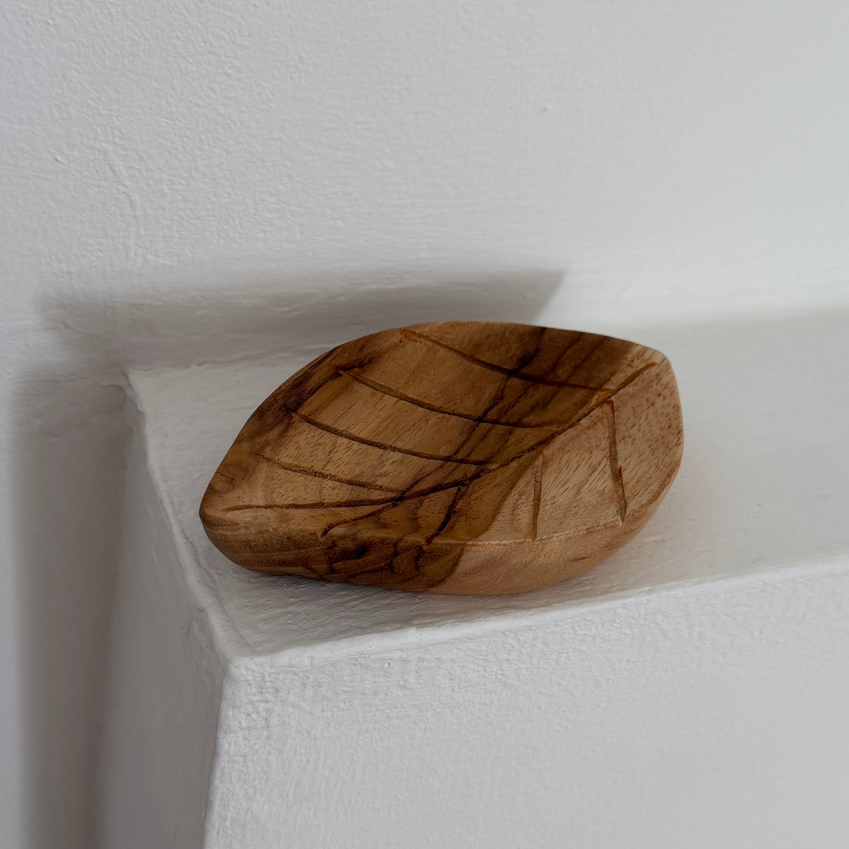 A small hand-carved wooden bowl shaped like a leaf, featuring natural dark grain lines, resting on a white textured shelf against a white wall.