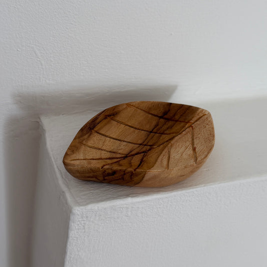 A small hand-carved wooden bowl shaped like a leaf, featuring natural dark grain lines, resting on a white textured shelf against a white wall.